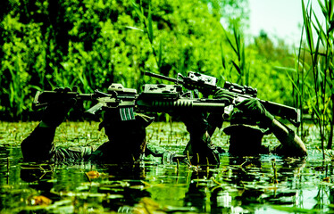 Two soldiers in camouflage gear and helmets wade through swamp water holding rifles with attached...