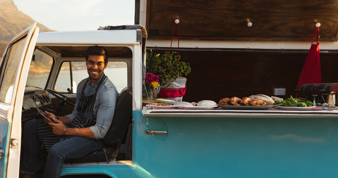 Hispanic man sitting in driver seat of turquoise van by waterfront, holding tablet, copy space