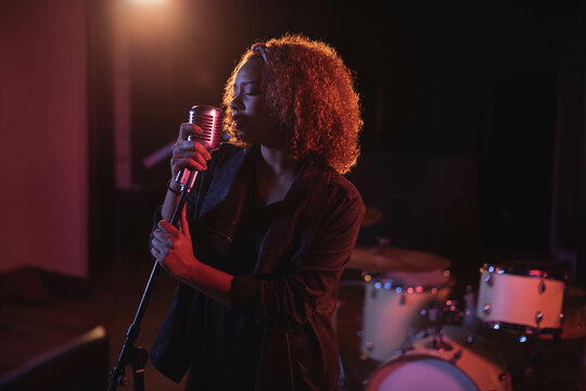 Singer standing on stage under red, purple lights holding vintage microphone with drum kit behind