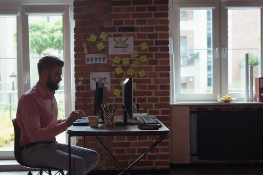 Bearded man in pink shirt working in brick office using dual monitors and sticky-notes, copy space