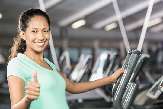 Woman in sportswear giving thumbs-up and standing on cardio machine at gym, showing ellipticals