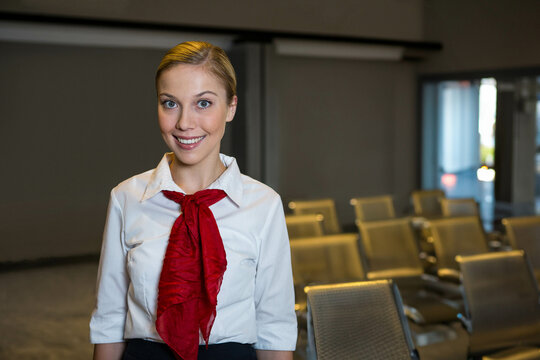 Female airline staff standing in gate lounge by metal seating, glass doors, signage rail and tiles