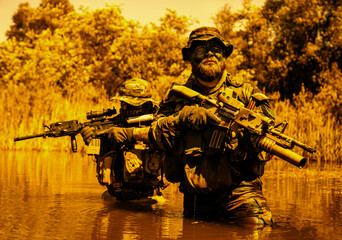 Two armed soldiers in camouflage and tactical gear wade through water, holding rifles, with trees and foliage in the background.