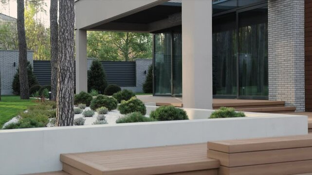 Slow motion shot of a garden terrace near a modern house. Modern residential design with landscaped garden, large windows and elegant wooden terrace.