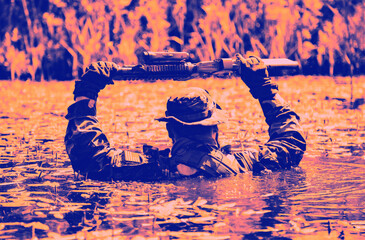 A soldier wearing camouflage holds a firearm above his head while wading through water surrounded by reeds in a dramatic color filter.