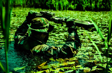 Soldier in camouflage wades through a swamp holding a rifle above the water surface surrounded by aquatic plants and greenery.