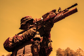 Soldier in camouflage attire and field helmet aiming a rifle with mounted optic under a yellow-orange sky, with trees in the background.