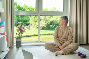  An elderly woman in a tracksuit sits on a rug at the big window in  the home in front of a laptop...