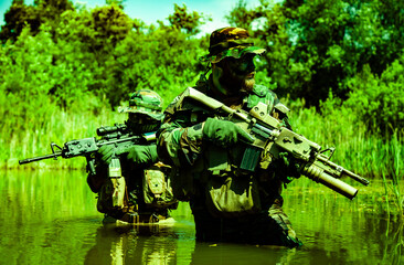 Two armed soldiers in camouflage with rifles standing in shallow water, surrounded by dense...