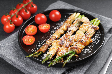 Grilled asparagus wrapped in crispy bacon sprinkled with parmesan cheese served on plate with fresh cherry tomatoes on black background. Gourmet dish, cooking recipe, cuisine, or restaurant menu