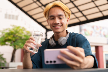 Asian gay teenager drinking water and using phone outdoors in cafe restaurant