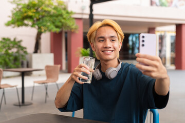 Asian gay teenager drinking water and using phone outdoors in cafe restaurant