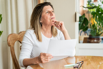 Senior woman sitting at dining table, holding documents and looking pensive, contemplating her...