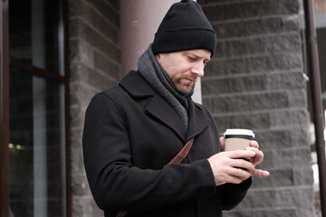 Pensive hipster drinking coffee in the urban street. Handsome man wearing black coat, brown leather...