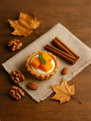 Fototapeta premium Autumn-themed tartlet with pumpkin cubes and mint on a rustic cloth, surrounded by cinnamon sticks, walnuts, pecans, and dry maple leaves. Cozy fall dessert composition.