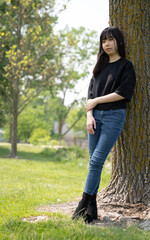 Young asian teenage girl standing on the ground in a grassy area, leaning on a tree, wearing a black top and jeans.