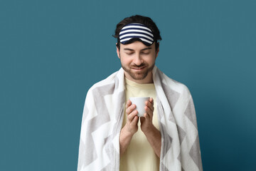 Handsome young man with sleep mask, cup of coffee and blanket on blue background
