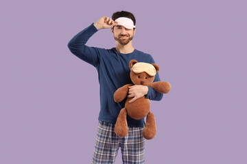 Handsome young man with sleep mask and teddy bear on lilac background