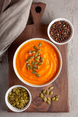 A bowl of vibrant orange pumpkin soup topped with pumpkin seeds, placed on a wooden board alongside bowls of whole peppercorns and raw seeds, creating a rustic and cozy food styling.