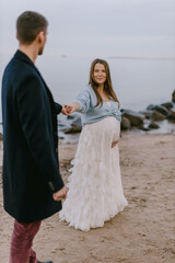 Happy pregnant woman holding partner’s hand on beach, smiling at camera. Maternity love, outdoor lifestyle and family expecting baby