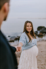 Happy pregnant woman holding partner’s hand on beach, smiling at camera. Maternity love, outdoor lifestyle and family expecting baby