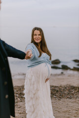 Happy pregnant woman holding partner’s hand on beach, smiling at camera. Maternity love, outdoor lifestyle and family expecting baby