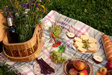 A cozy picnic setting with a wicker basket filled with wildflowers and wine, cheese on bread, fresh pears, apples, grapes, and cured meats, all arranged on a soft plaid blanket in the grass.