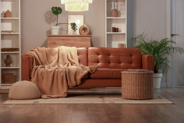 Interior of modern living room with brown sofa, wicker basket, blanket and glowing lamp at evening