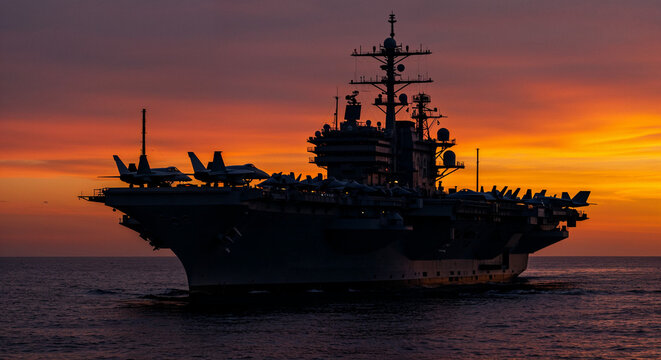 Silhouetted US Navy supercarrier at sunset with jets on deck and glowing ocean reflections.

