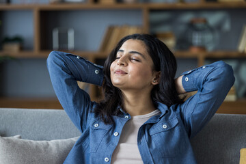 Close-up photo of a young Indian woman sitting on the couch at home, eyes closed, hands behind her...