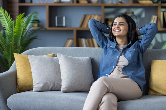 Smiling and relaxed young Indian girl sitting on the couch at home, hands behind her head and resting