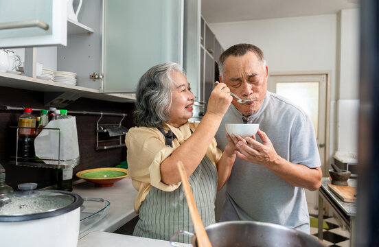 Happy Senior Asian Woman Feeding Her Husband in the Kitchen, Elderly Couple Enjoying a Home Cooked Meal Together, Mature Asian Spouses Tasting Food in Kitchen - Powered by Adobe