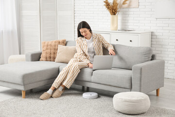 Young woman in pajamas using laptop on sofa with robot vacuum cleaner at home