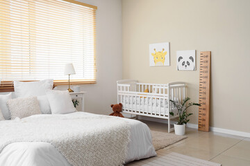 Interior of modern bedroom with big bed, baby's crib, paintings and wooden stadiometer near window