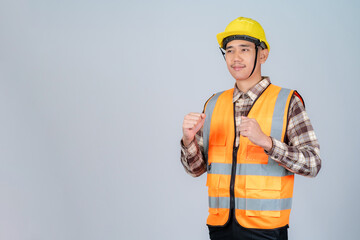 Young Asian male engineer or worker wearing yellow hard hat and reflective vest standing on grey background.