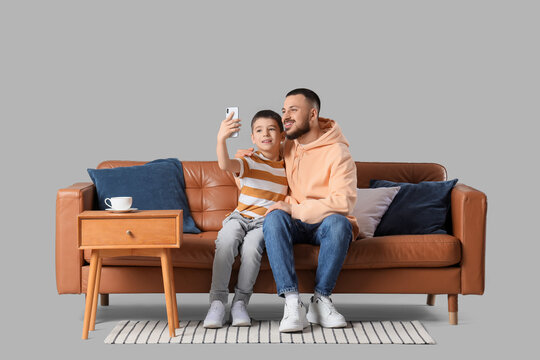 Happy father with his son taking selfie on brown leather sofa against grey background