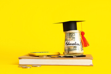 Graduation hat, book and jar with money for education on yellow background. Tuition fees concept