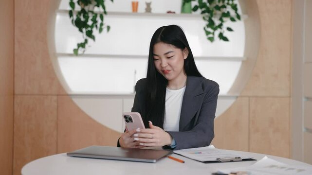 Delighted Asian businesswoman sitting at desk clenching fist with joy while looking at smartphone. Smiling with excitement after receiving great news. Celebrating success or milestone in workspace.
