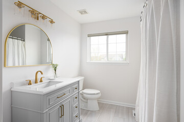 A bathroom with a grey cabinet, white countertop, tile flooring, and gold faucet, light fixture, and semicircle mirror.