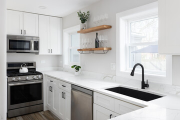 A white kitchen with a marble countertop, black faucet and sink, decorations on floating wood shelves, and stainless steel appliances.