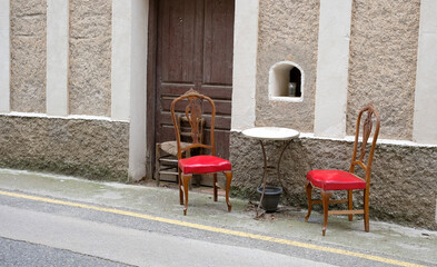 Two red chairs and a round table on a deserted street near a blank wall. A metaphorical view of...