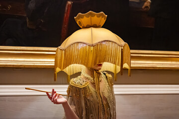 A surreal and theatrical figure stands in a gilded vintage gown and ornate lampshade headpiece,...