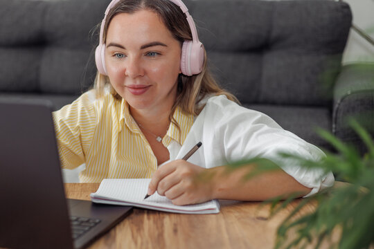 Young Caucasian woman with blond hair wearing pink headphones writing in notebook while using laptop at home desk