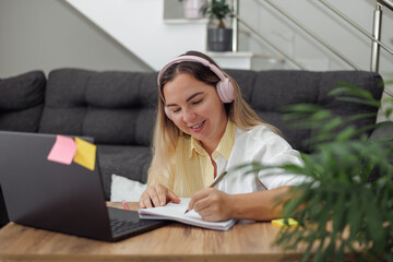 Young blond European woman with headphones sitting at wooden table, writing in notebook, using laptop, focused on work or study.