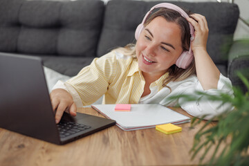 Young Caucasian woman with brown hair wearing headphones using laptop and writing in notebook at wooden table indoors.