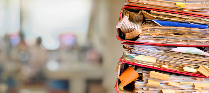 stack of messy file folders with narrow depth of field, blurred office in the back,beat the clock,stress,red tape, bureaucracy, heavy workload,business concept. 