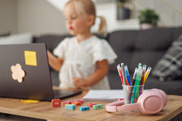 Blond European toddler girl with laptop and colorful pencils on wooden table in living room, early...
