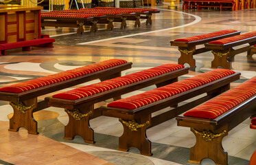 Rows of ornate church kneelers with red cushioned seats inside an elegant sanctuary. Decoratively...