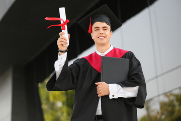 Male graduating student with diploma and book near university