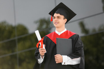 Male graduating student with diploma and book near university